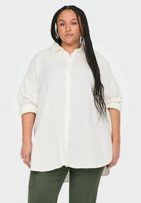 Chemise blanche à boutons en tissu léger, dotée d'un col pointu, de manches retroussées et d'un ourlet incurvé. Associée à un pantalon vert.