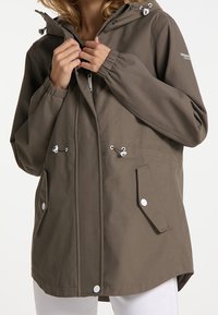 Veste marron résistante à l'eau avec capuche, fermeture éclair frontale, poignets élastiques et poches à boutons-pression. Dotée d'un cordon de serrage et de détails blancs.