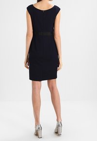 Mörkblå klänning med figurnära passform och korta ärmar, insvängd midja som betonas av ett svart bälte, och en dold dragkedja. Bärs med silverfärgade högklackade skor.