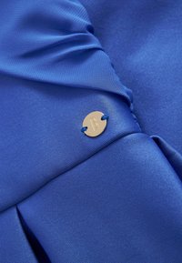 Sininen kangas, jossa on sileä tekstuuri, pleats ja pyöreä kultainen yksityiskohta, jossa on "B"-logo, ommeltu sinisellä langalla.