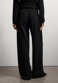 Zwarte, wijde broek met een gladde textuur, voorzien van twee achterzakken en een getailleerde pasvorm, gedragen met een langemouwen top.