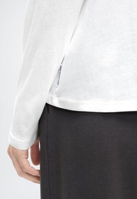 Χέρι ακουμπισμένο στο πλάι, φορώντας λευκό πουκάμισο με μακριά μανίκια που έχει ετικέτα από ύφασμα και μαύρο παντελόνι.