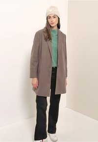 Bruine wolmix jas met een knoopsluiting, twee zakken, gedragen over een groene trui en zwarte flare-broek, gecombineerd met een beige gebreide muts.