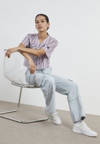 Violetā, svītrainā, apgriezta džersija kombinācijā ar gaiši ziliem plašiem džinsiem un baltām kedām; modelis sēž uz caurspīdīgas krēsla.