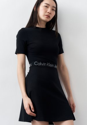 Femeie purtând o rochie neagră cu mâneci scurte, cu logo-ul "Calvin Klein Jeans" pe talie, stând în fața unui perete simplu, de culoare deschisă.