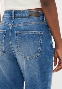 Casaco de ganga azul com uma cintura alta, apresentando um patch de marca em pele e dois bolsos traseiros, mostrando um efeito de lavagem desbotada e detalhes de costura.
