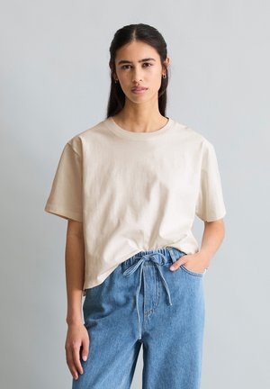 Kobieta o długich ciemnych włosach nosi luźną beżową koszulkę wsuniętą w niebieskie jeansy z wysokim stanem i sznurkiem w pasie, stojąca na jednolitym tle.