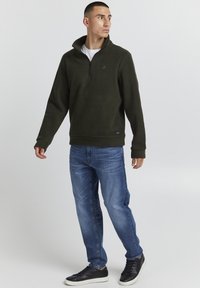 Pullover in pile verde scuro con zip a un quarto e colletto alto, abbinato a jeans blu e sneakers nere, con un logo discreto sul petto.