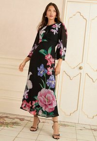 Čierne maxi šaty s kvetinovým vzorom, ktorý zdobia farebné pivónie a iris. Dlhé priesvitné rukávy a priama silueta. Ozdobené jemnou textúrou.