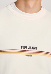 Camiseta de algodón en color claro con cuello redondo. Presenta el texto "PEPE JEANS" y "LONDON W11" impreso en negro, con acentos de rayas horizontales en amarillo, naranja y rojo.