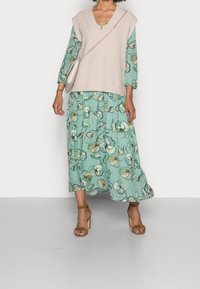 Mujer vestida con un vestido midi floral color teal, un chaleco sin mangas de punto beige, tacones con correa al tobillo color marrón y un bolso cruzado beige con cadena dorada.