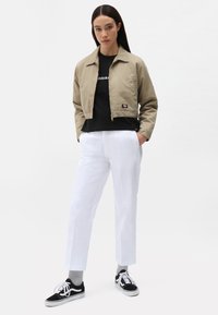 Beige, kort jakke med glidelås, svart grafisk t-skjorte, hvite rettskåret bukser og svarte joggesko med hvite detaljer, mot en lys bakgrunn.