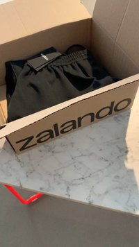 Črne kratke hlače v kartonski škatli z blagovno znamko Zalando. Hlače imajo elastičen pas in oznako na vrhu, лежаčne на мрамору.