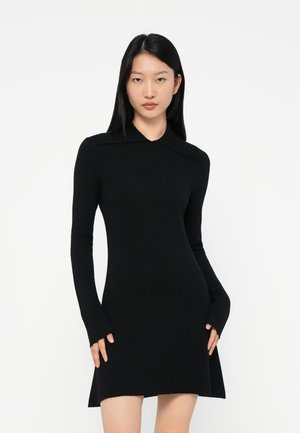 Young woman with straight black hair wearing a fitted long-sleeve black dress with a small collar and slightly flared hem, standing against white background.