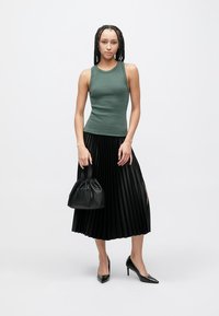 Grünes eng anliegendes Tanktop kombiniert mit einem schwarzen plissierten Midirock, schwarzer Handtasche und spitzen High Heels; durchgehend glatte Textur.