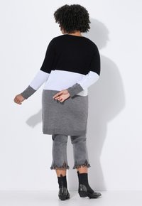Person mit lockigem Haar, der einen langen, farblich abgestimmten Pullover, ausgefranste graue 3/4-Hosen und schwarze, mit Nieten besetzte Stiefeletten trägt und mit dem Rücken zur Kamera steht.