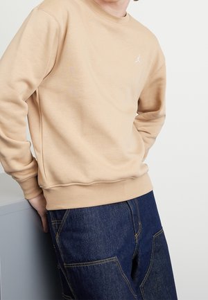 Person iført en beige sweatshirt med en lille hvid logo på brystet og mørkeblå denimbukser, hvilende på en hvid overflade.