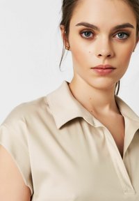 Beige Poloshirt mit einem Kragen und kurzen Ärmeln. Glatte Textur; verfügt über einen subtilen Glanz und saubere Nähte.