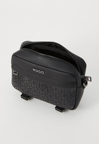 Black synthetic bag with embossed "HUGO" pattern, smooth top, dual zippers, and adjustable straps. Compact rectangular design.
