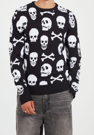 Pull noir avec motifs blancs de têtes de mort et os croisés, porté avec un jean gris délavé, montrant le haut du corps et une main.