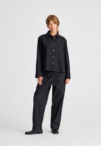 Cămășă oversized din bumbac negru cu nasturi și guler ascuțit, combinată cu pantaloni largi asortați și pantofi plate negri.