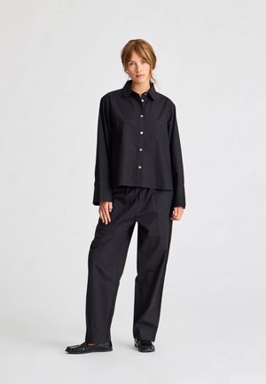 Camicia oversized in cotone nero con bottoni e colletto a punta, abbinata a pantaloni larghi coordinati e scarpe basse nere.
