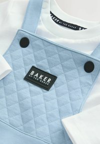 Svetlo modri kviltani kombinezoni z nastavljivimi naramnicami, ki vsebujejo črno oznako "BAKER". Pod njimi se vidi bela majica.