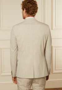 Chaqueta de traje beige claro con una tela texturizada, corte ajustado, abertura simple en la parte posterior y adornos de cuatro botones en los puños.