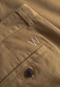 Pantalon kaki en tissu texturé, avec des coutures détaillées, un bouton de taille visible et un logo brodé sur la poche.