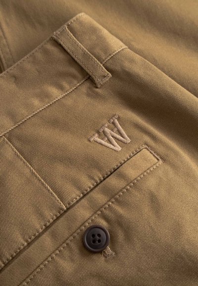 Pantalon kaki en tissu texturé, avec des coutures détaillées, un bouton de taille visible et un logo brodé sur la poche.