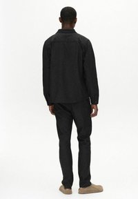 Black denim shirt and pants with a textured finish. Shirt features a collar and button cuffs. Model is wearing tan slip-on shoes.