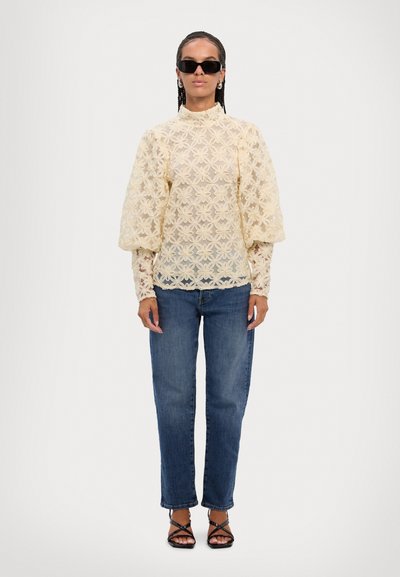 Blonde bluse med blomsterdesign i lys gul, høy hals og pufferm, parret med blå jeans og svarte sandaletter med stropper.