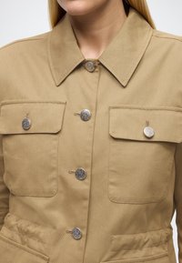 Chaki spalvos striukė su saga užsegimu, dviem krūtinės kišenėmis, sidabrinės spalvos sagomis ir struktūruotu apykakliu. Medžiaga yra lygi, šiek tiek reljefiška.