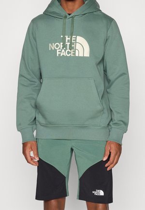 Mężczyzna w zielonej bluzie z kapturem North Face z przednią kieszenią oraz pasujących zielono-czarnych szortach North Face.