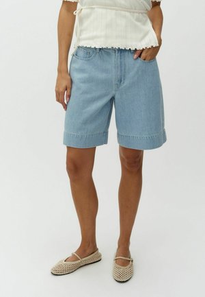 Persoon draagt lichtblauwe denim knielange shorts, wit gebreid topje met zijstrik en beige mesh ballerina's, staand tegen een effen achtergrond.