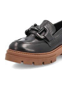Mocassin en cuir noir avec semelle épaisse en caoutchouc marron et grosse boucle décorative sur le dessus, vue en angle montrant les détails de l'avant et du côté.