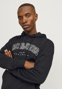 Sweat à capuche noir avec impression blanche "ZACK & JONES EST. 90 WORLDWIDE." Dispose d'une capuche à cordon et de poignets côtelés. Tissu doux et texturé.