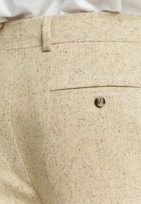 Pantalons beige en chevrons en tissu texturé, présentant une poche arrière avec un bouton et un motif légèrement chiné.