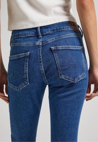 Los vaqueros de mezclilla azul tienen un diseño ajustado, estilo de cinco bolsillos y costuras curvas en los bolsillos traseros. Se puede ver la textura de la tela.