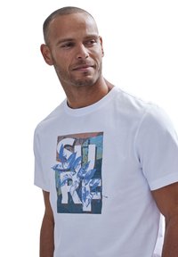 Weißes T-Shirt mit kurzen Ärmeln, das mit einem bunten grafischen Druck verziert ist, der blaue Blätter und abstrahierte Formen in erdigen Farbtönen zeigt. Weicher Baumwollstoff.