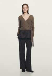 Maglione marrone a maniche lunghe con scollatura a V profonda, indossato sopra una maglietta di satin scuro, abbinato a pantaloni a righe con vita alta e scarpe nere.