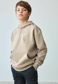 Bež dukserica s prednjim džepom, na kojoj je bijeli natpis "BAKER BY TED BAKER." Ima opušteni kroj i duge rukave.
