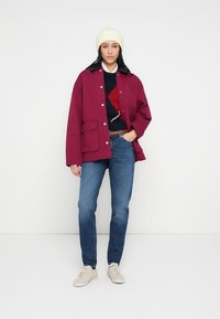 Bordowa, oversize kurtka z przednimi kieszeniami i zapinana na guziki, zestawiona z ciemnym swetrem ozdobionym czerwonym wzorem, niebieskimi dżinsami i białymi trampkami.