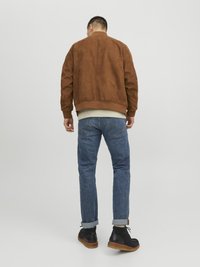 Chaqueta bomber de ante marrón con puños acanalados, sobre un fondo claro, combinada con jeans azules y botas negras de tobillo.