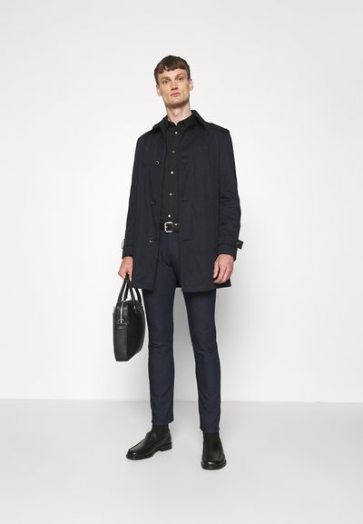 Svart trenchcoat med knappar, bärs över en svart skjorta. Mörkblå slim-fit byxor och svarta chelsea boots. Håller en svart läderväska.
