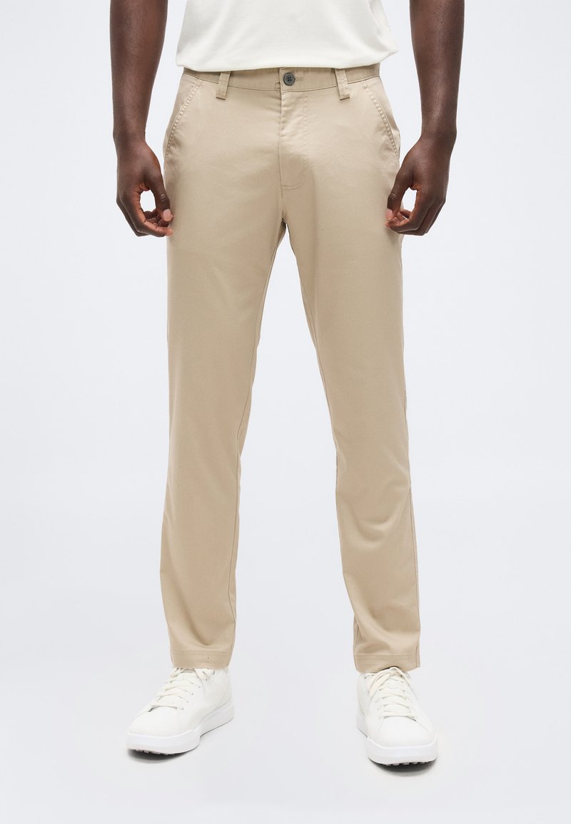 Άνδρας που φοράει μπεζ παντελόνι με ίσια γραμμή και πλαϊνές τσέπες, καθώς και λευκά αθλητικά, στέκεται μπροστά σε απλό φόντο.