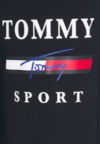 Schwarzes Sportshirt mit fettem weißen Text "TOMMY" und "SPORT", verziert mit einem bunten Streifen und einer Unterschrift in Blau und Rot.