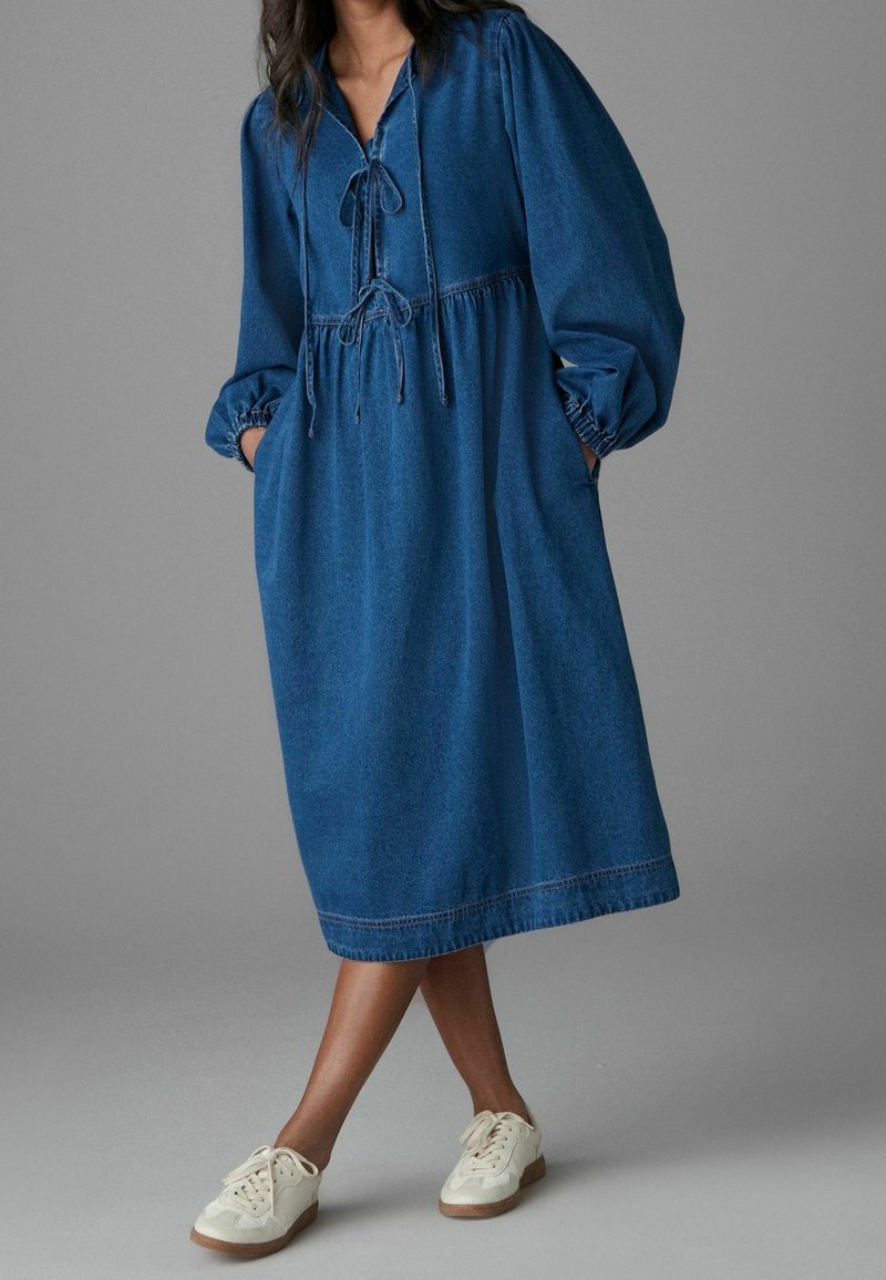 Robe en denim bleu à coupe ample, col à cordon, manches bouffantes et poches latérales; longueur mi-mollet avec un design décontracté.