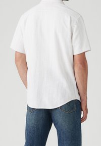 Chemise blanche à manches courtes avec des rayures verticales fines. Dotée d'un col classique et d'un ourlet arrondi. Tissu texturé, associée à un jean en denim bleu.