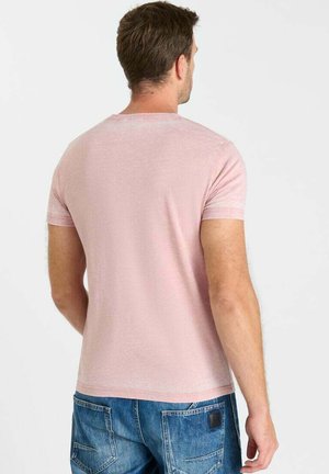 Man die een lichtroze T-shirt met korte mouwen en een blauwe spijkerbroek draagt, van achteren gezien tegen een effen lichte achtergrond.